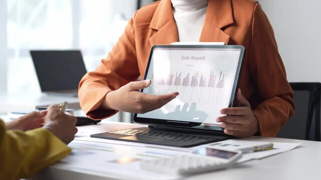 A woman in an orange jacket is looking at a computer screen with a graph on it. She is pointing at the graph and seems to be explaining something to someone else. Concept of collaboration and learning