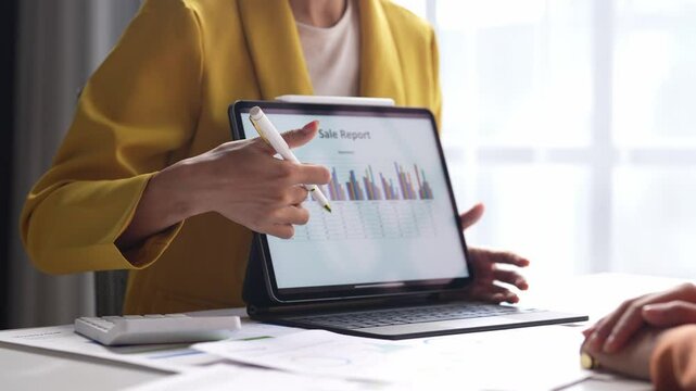 A woman is showing a laptop to another person. The laptop screen displays a sales report. The woman is pointing to the report and explaining it to the other person