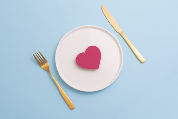 Top view of paper heart on white plate with golden cutlery on pastel blue background. Minimal Valentine's Day dinner invitation card, top view