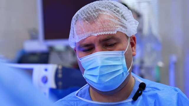 Face of a male Caucasian doctor in cap and mask. Female scrub nurse appears in the frame. Close up. Blurred backdrop.