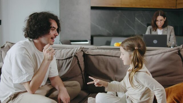 Loving curly haired father and daughter playing together on the couch at home, while business mom works in the kitchen.