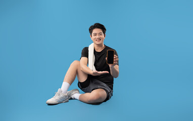 Full body image of Asian guy wearing gym clothes, using phone and posing on blue background