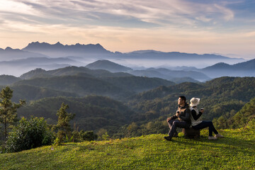 Asian man and woman campers sitting on rock and having morning coffee enjoy mountains view