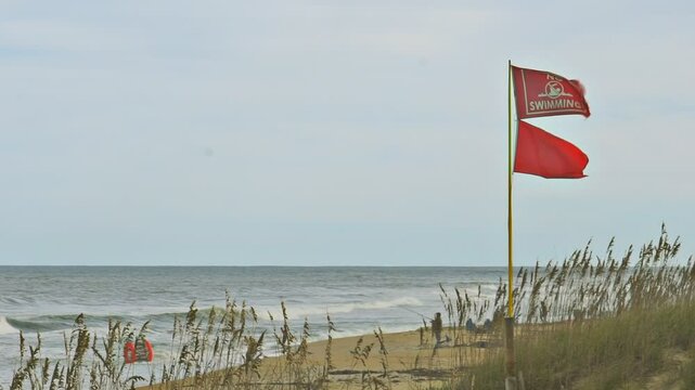 Beach warning flags, the red flag, indicating high hazard/ and or strong currents blows in the breeze with a red "no swimming" flag on the shore with some people ignoring the warning.