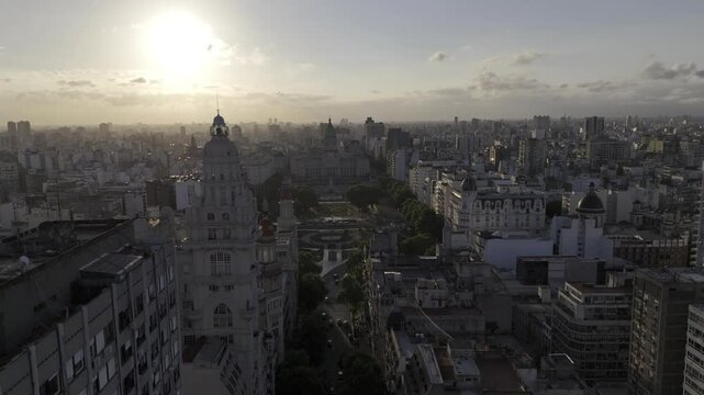 Drone flies down Avenida de Mayo, past Palacio Barolo and over Plaza Mariano Moreno toward the National Congress with a sunset behind it in Buenos Aires, Argentina