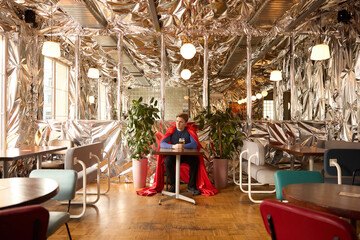Superhero in bright cape sits in futuristic cafe showcasing pause in heroic duties to embrace leisure, connecting fantasy with human simplicity. Concept of future and past, life-work balance, relax.
