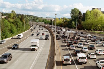 A busy freeway in the United States. Traffic is congested with cars, trucks, and vans moving slowly. The road is lined with trees and buildings, suggesting a suburban or urban area. The sky is blue. © Apichaya