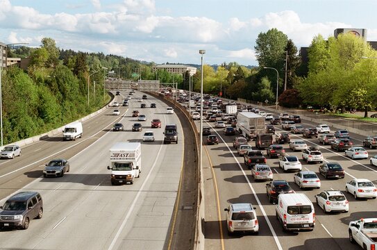 A busy freeway in the United States. Traffic is congested with cars, trucks, and vans moving slowly. The road is lined with trees and buildings, suggesting a suburban or urban area. The sky is blue.