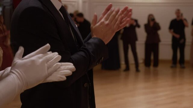 Man in suit and young woman in luxurious gown and gloves are standing and applauding at festive event in honor of special occasion.