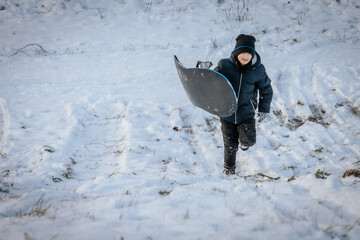 A boy dressed in a black winter jacket and hat carries a sled while walking uphill on a...