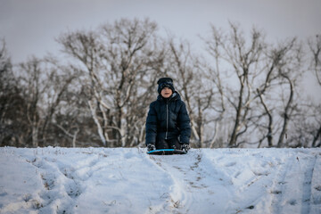 A smiling boy in a black winter jacket and hat sleds downhill on a snow-covered slope. Bare trees...