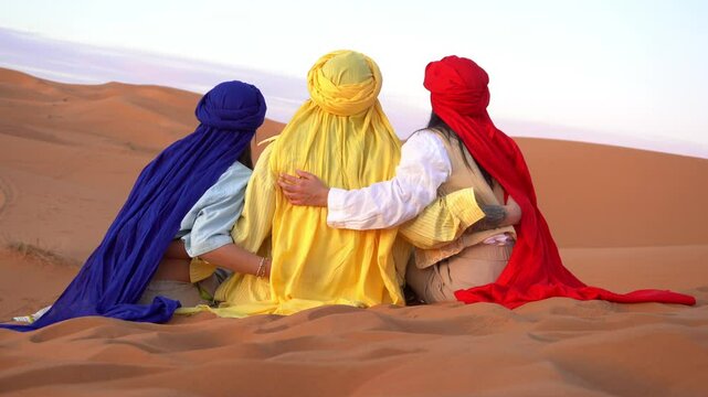three islamic tourist arab women with colorful red, yellow and blue turbans sit in the Sahara desert in Morocco Africa watching the sand dunes and sunset - trip to  Merzouga to visit African nature