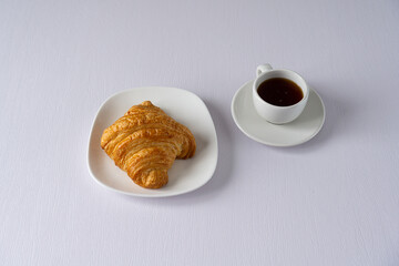 A simple breakfast setup featuring a freshly baked croissant on a white plate and a cup of black coffee on a white table, creating a clean and minimalistic presentation.
