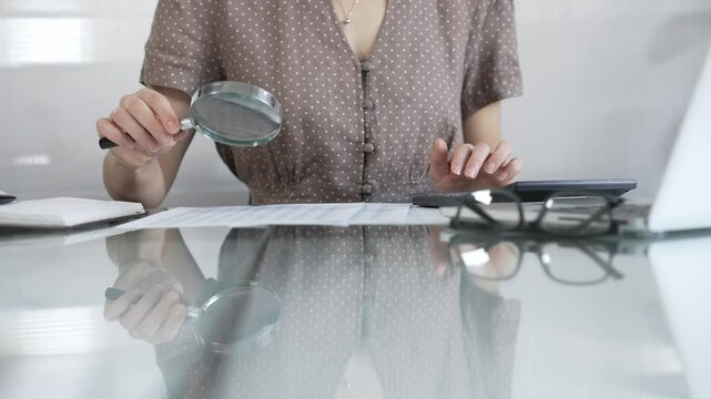 Businesswoman accountant in brawn dotted blouse reviewing financial reports with magnifying glass and calculator, conducting audit and analyzing data in office. Taxes, audit in business