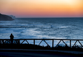 Fototapeta premium Silhouette of a person admiring the ocean waves during a vibrant sunset over the Ericeira coast.