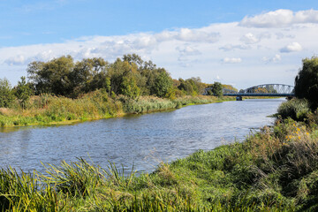 Scenic river landscape. Distant bridge over the water. Surrounded by lush greenery and wild nature. Water reflecting sky. Riverbank environment. Summer holiday by the water. Wide river flowing.