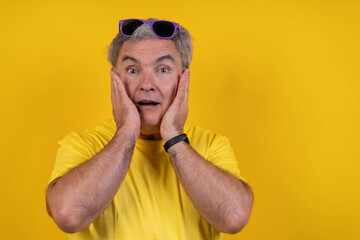 Surprised mature man with sunglasses on head holding cheeks with hands on yellow background