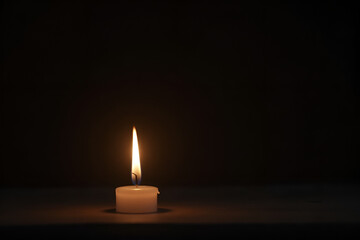 Single burning candle flame or light glowing on a small white candle on black or dark background on table in church for Christmas, funeral or memorial service with copy space