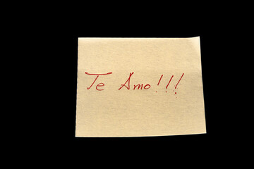 Yellow adhesive paper, similar to a Post-it note, on a black background with the words “Te Amo” in Spanish handwritten in red ink.