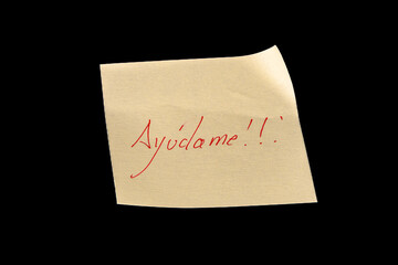 Yellow adhesive paper, similar to a Post-it note, on a black background with the words “Ayúdame” in Spanish handwritten in red ink.
