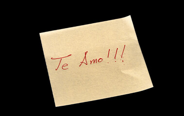 Yellow adhesive paper, similar to a Post-it note, on a black background with the words “I love You” “Te amo” in Spanish handwritten in red ink.