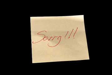 Yellow adhesive paper, similar to a Post-it note, on a black background with the word “Sorry” and three exclamation marks handwritten in red ink.