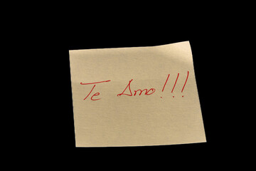 Yellow adhesive paper, similar to a Post-it note, on a black background with the words “Te Amo” in Spanish handwritten in red ink.