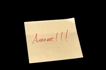 Yellow adhesive paper, similar to a Post-it note, on a black background with the words “Amame” “Love me” in Spanish handwritten in red ink.