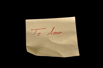 Yellow adhesive paper, similar to a Post-it note, on a black background with the words “I love You” “Te amo” in Spanish handwritten in red ink.