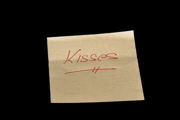 Yellow adhesive paper, similar to a Post-it note, on a black background with the word “Kisses”  handwritten in red ink.