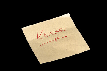Yellow adhesive paper, similar to a Post-it note, on a black background with the words “Kisses” handwritten in red ink.