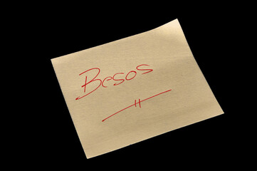 Yellow adhesive paper, similar to a Post-it note, on a black background with the words in Spanish “Besos”  handwritten in red ink.