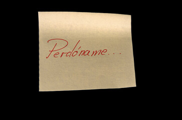 Yellow adhesive paper, similar to a Post-it note, on a black background with the word “Perdóname”  in Spanish handwritten in red ink.
