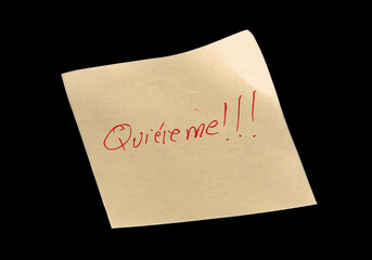 Yellow adhesive paper, similar to a Post-it note, on a black background with the words “Quiéreme” “Love me” in Spanish handwritten in red ink.