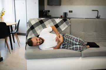 Handsome young man suffering from stomach ache while lying on sofa at home