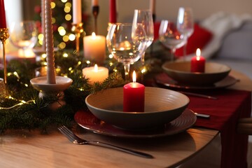 Beautiful dishware with burning candles, fir tree branches and Christmas lights on wooden table indoors, closeup