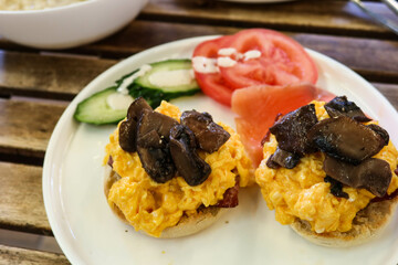 close up of American breakfast with scramble egg on muffin bread in half and stir fried mushroom. uncooked tomato and cucumber on the side on a white plate on a wooden table