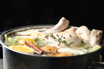 Whole fresh turkey, cut citrus fruits, thyme and spices boiling in pot against black background, closeup