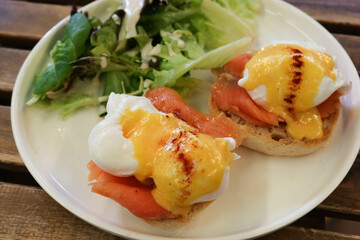 Close up of English breakfast of egg Benedict with salmon and cream muffin bread. Salad vegetables on the side on a white plate on a wooden table (egg royale)