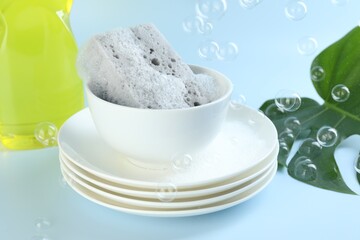 Sponge with foam and stack of dishware among flying bubbles on light blue background, closeup