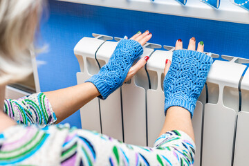 girl in knitted mittens warms her hands on a radiator.