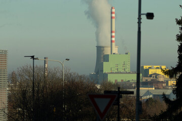 A large power plant with tall smokestacks emitting smoke, located in a busy urban area with many...