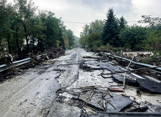 Destruction on a rural road after a natural disaster. Flood damage with debris, broken asphalt, and fallen trees under a cloudy sky. A somber scene highlighting the impact of climate change.