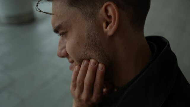Close-up face of frustrated sick ill man touching cheek sitting on bench, gripping face in discomfort enduring painful toothache. Male closing eyes with expression of suffer from painful toothache