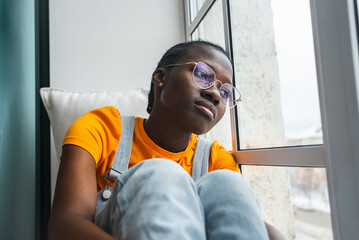 Pensive young African American woman with glasses is sitting by the window, embracing her knees and...