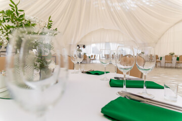 Close up of wine glasses, silver tableware and green napkins on round tables covered with white tablecloth.