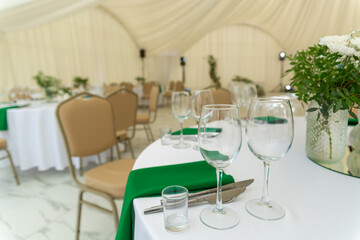 Close up of wine glasses, silver tableware and green napkins on round tables covered with white tablecloth.