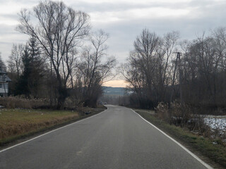 A serene road surrounded by leafless trees under a cloudy sky, leading into the horizon with a hint of sunlight peeking through the clouds