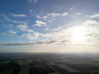 Aerial view of a vast rural landscape with patchy fields under a cloudy sky. The sunlight creates a warm glow, contrasting with the cool tones of the fields.