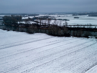 Aerial view of a snowy landscape with frozen river and bare trees, creating a serene winter scene.